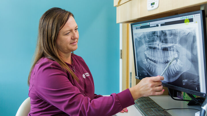 Woman with long brown hair is wearing purple scrubs and points at an xray of teeth with a gray pen