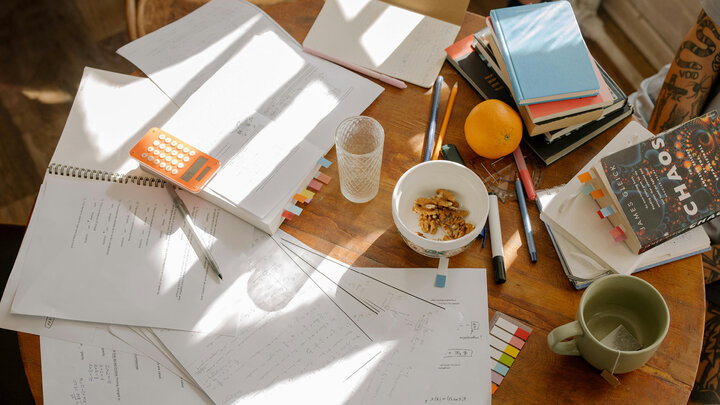 A cluttered wooden table with papers, notebooks, a calculator, pens, an orange, a bowl of nuts, a glass of water, a green mug, and a book titled “Chaos.”