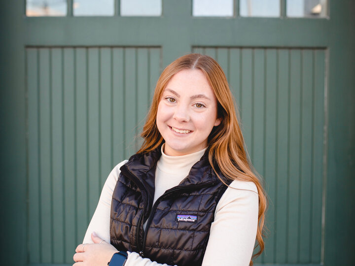 Women with light brown hair standing in front of a green door. She is wearing a white long sleeve shirt and black vest.
